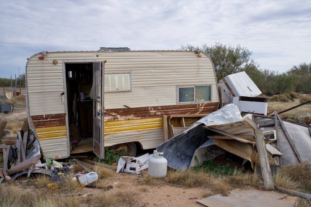 A weathered abandoned trailer surrounded by debris and trash in a rural outdoor setting.