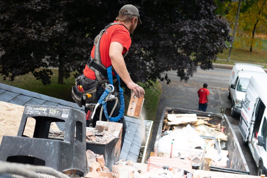 Worker in safety harness removes bricks from roof into dumpster below.
